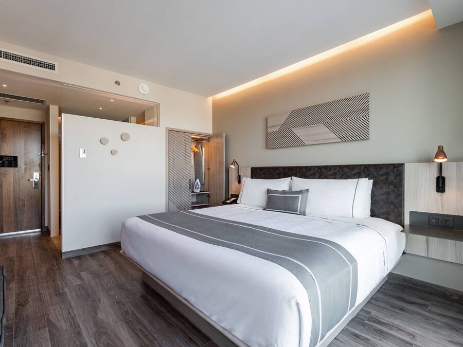 Quarto de hotel moderno com uma cama grande, um guarda-roupa de madeira, um espelho e uma luminária fixada na parede.