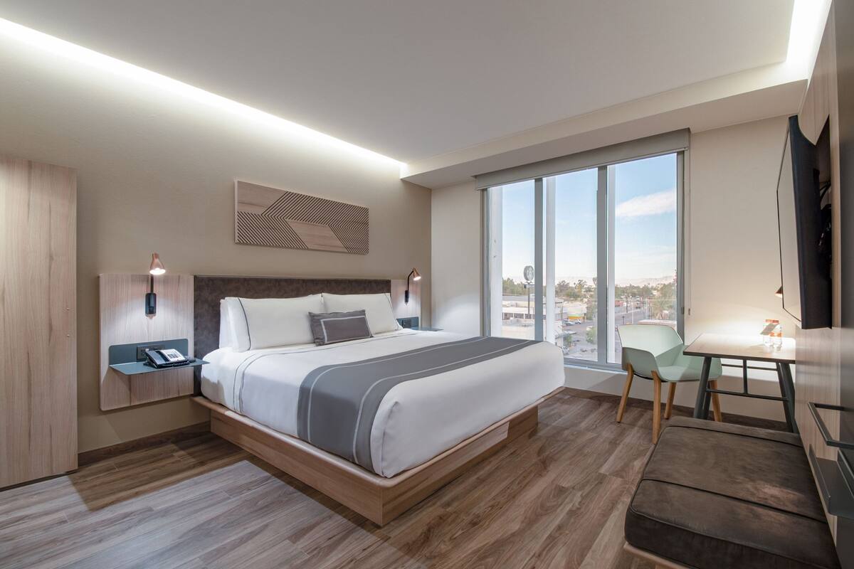 Ein modernes Hotelzimmer mit einem großen Bett, einem Schreibtisch und Blick auf die Stadt durch große Fenster.