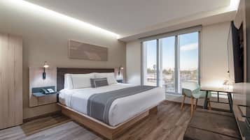A modern hotel room with a large bed, a desk, and a view of the city through large windows.