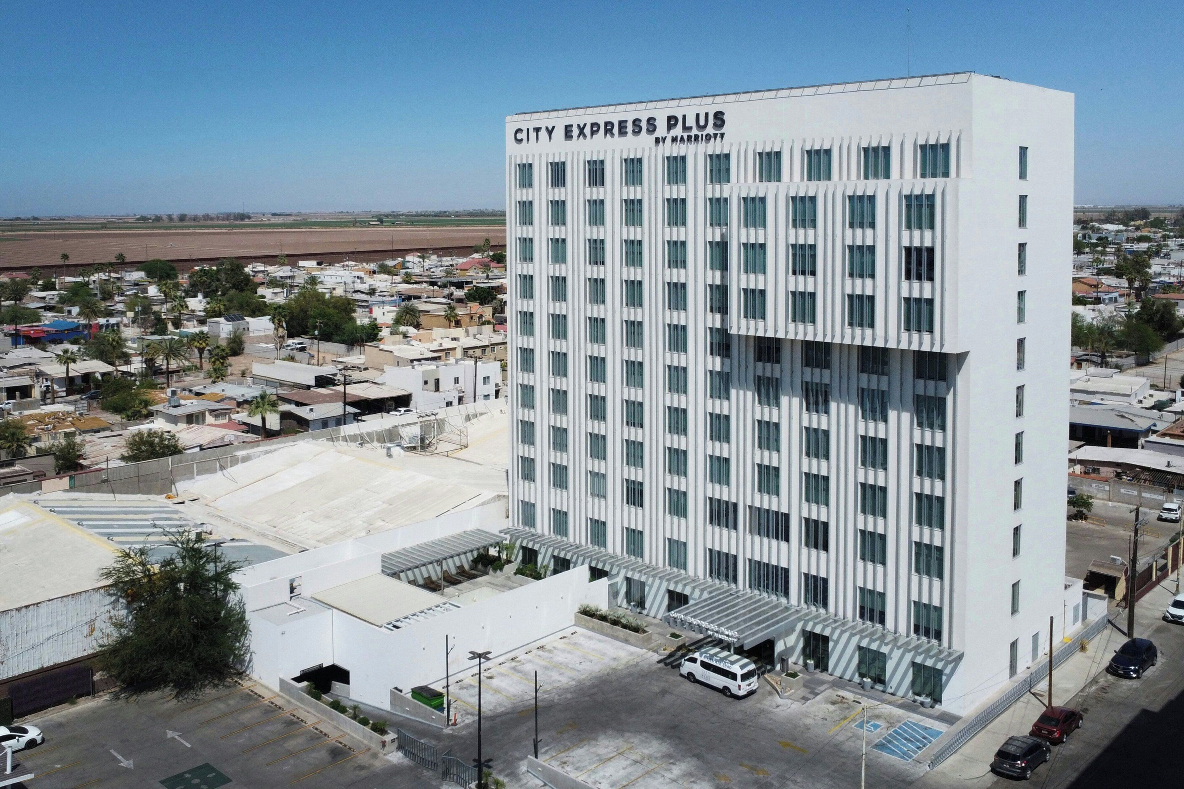 A modern hotel with a prominent 'CITY EXPRESS PLUS' sign, a parking lot, and a view of the surrounding urban area.