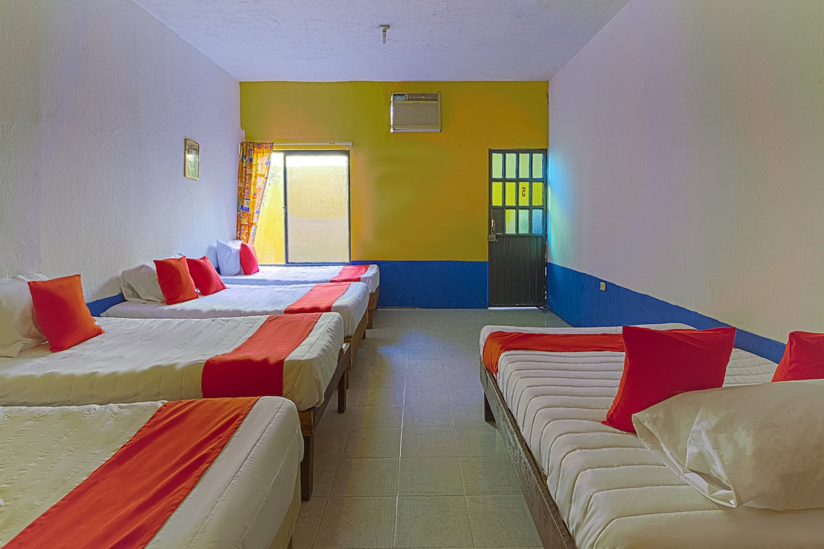 A hotel room with four beds arranged in a row, a window with curtains, a door with a window, and a ventilation unit on the wall.
