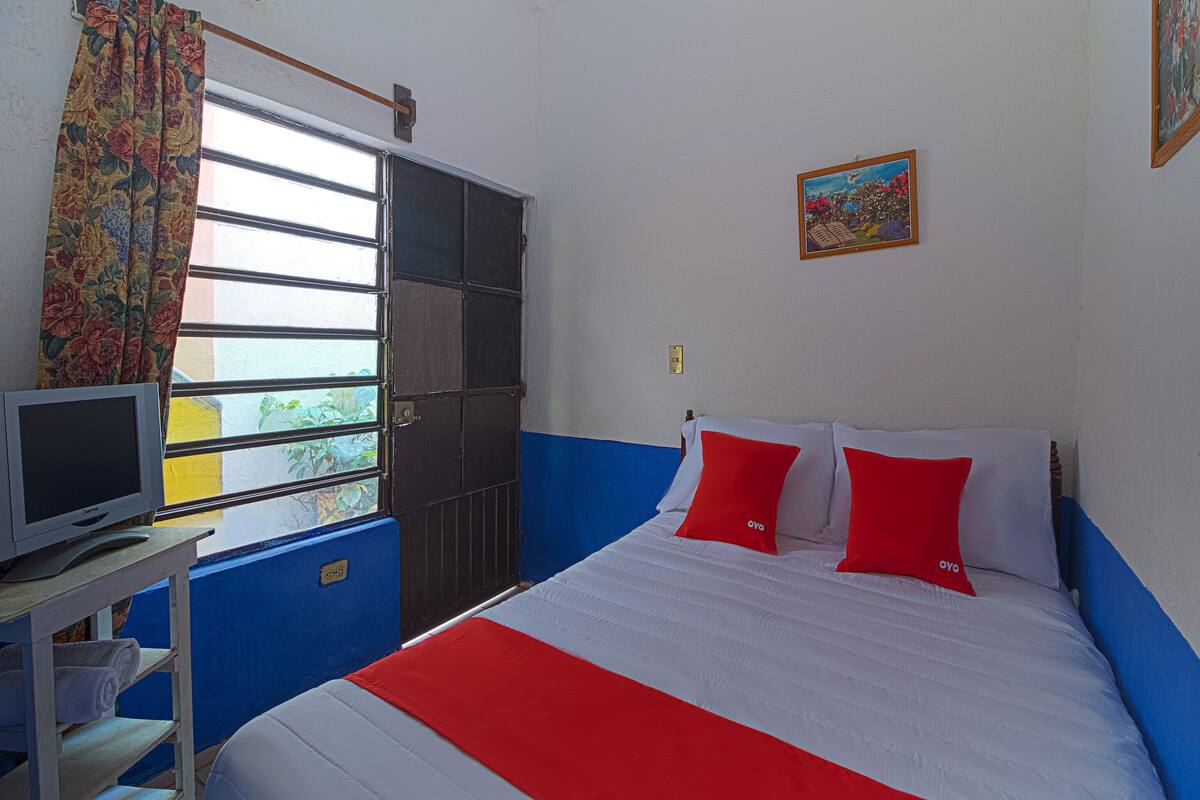 A room with a bed, a television on a stand, a window with curtains, and a painting on the wall.