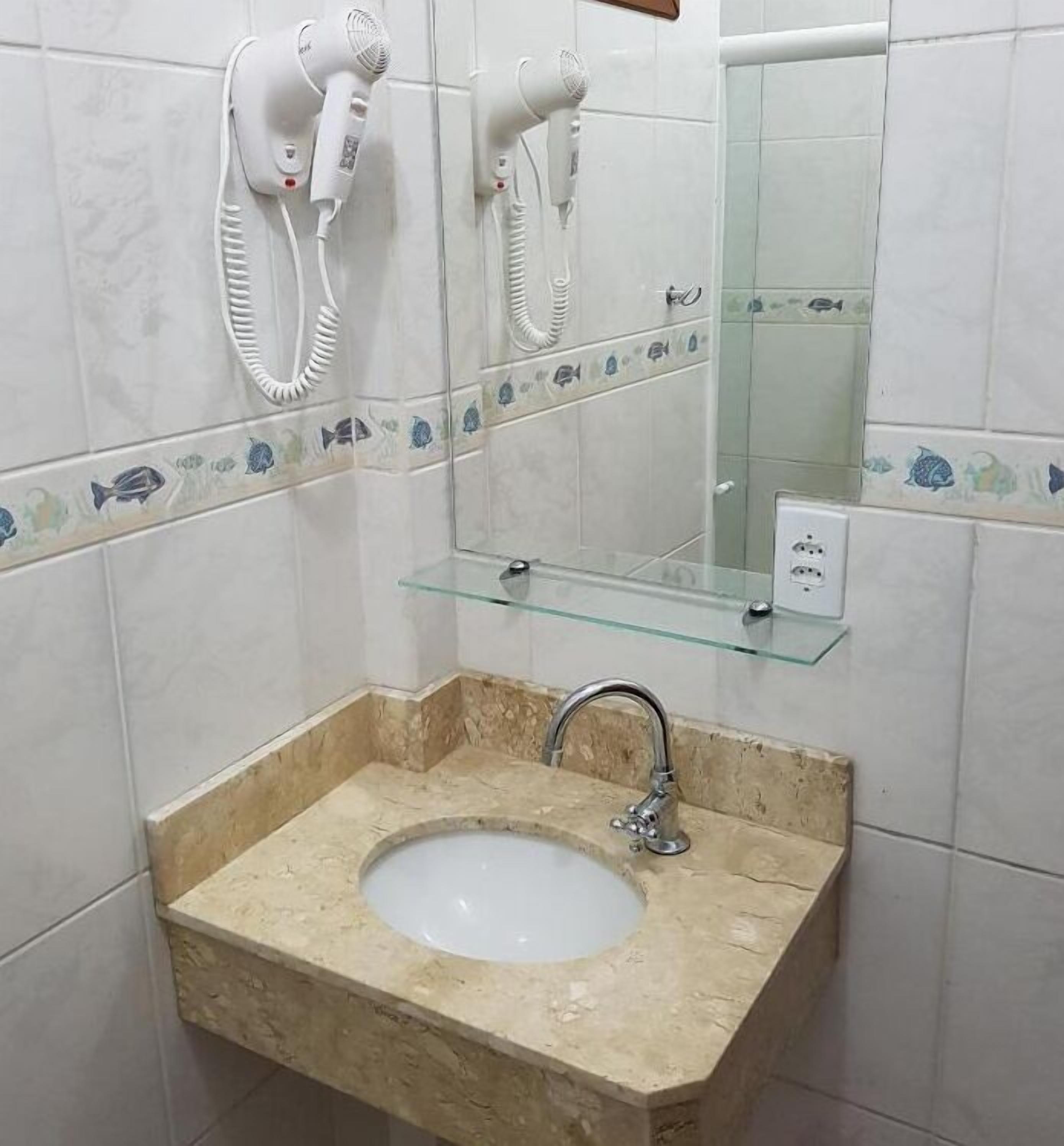 Une salle de bain avec un lavabo beige, une étagère en verre et deux sèche-cheveux fixés au mur.