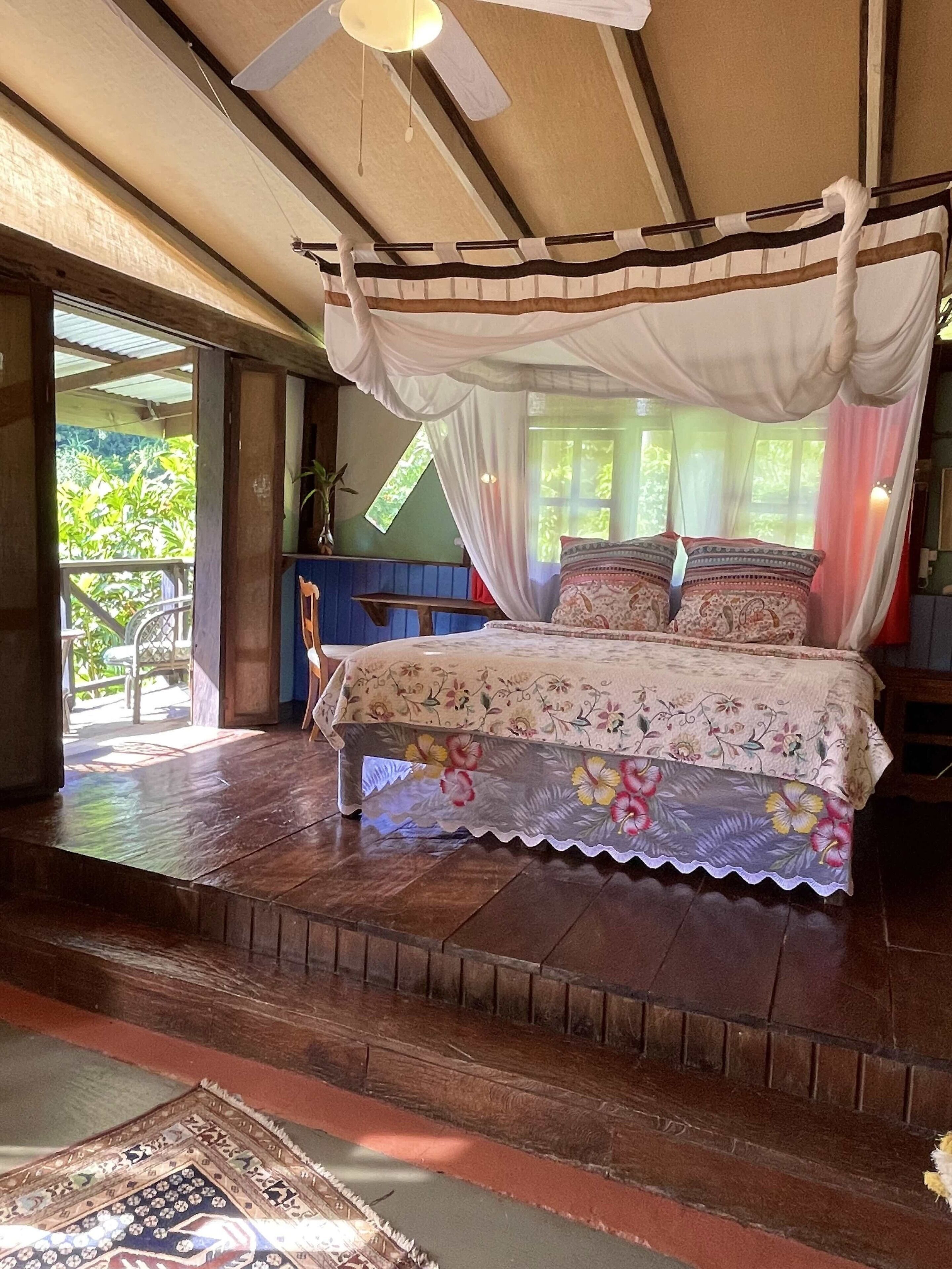 A room with a bed, a ceiling fan, a window with curtains, and a view of greenery outside.