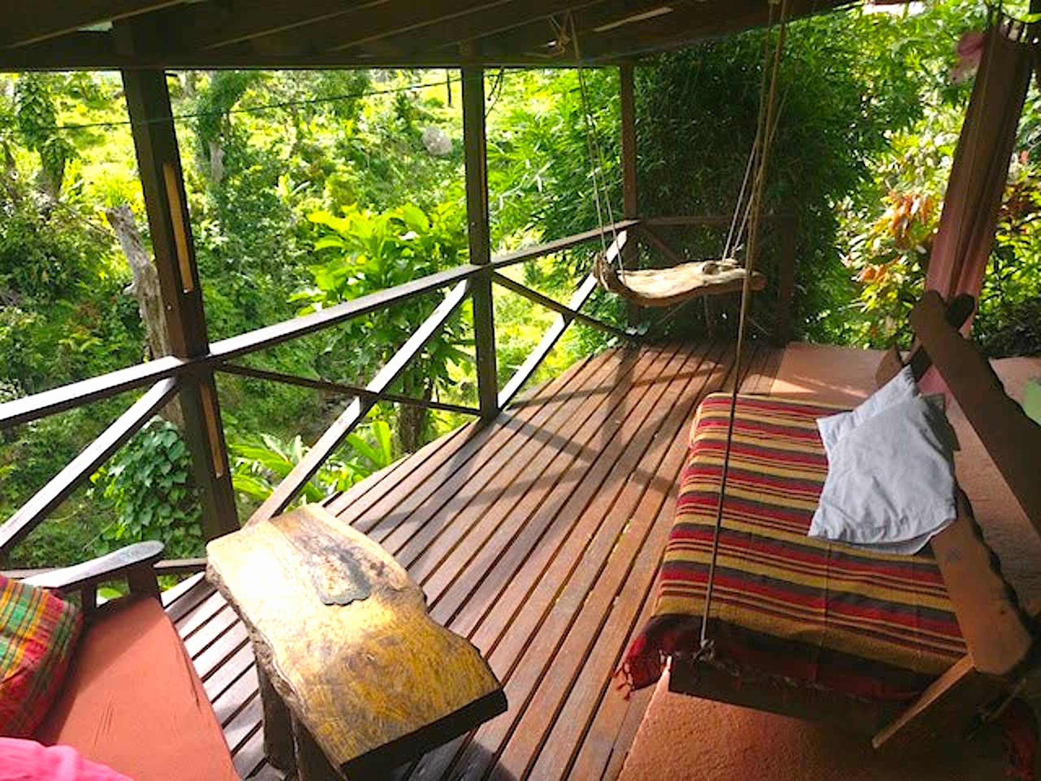 Una terraza de madera con una hamaca y un banco acolchado, con vistas a un exuberante bosque verde.