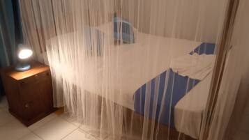 A bed with a white and blue bedspread, a bedside table with a lamp, and a sheer curtain.
