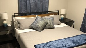 Una cama bien tendida con cubrecama acolchado, dos almohadas, una manta azul y un cabecero. Se ven dos mesitas de noche con lámparas, una alfombra oscura y una cortina azul en la ventana.