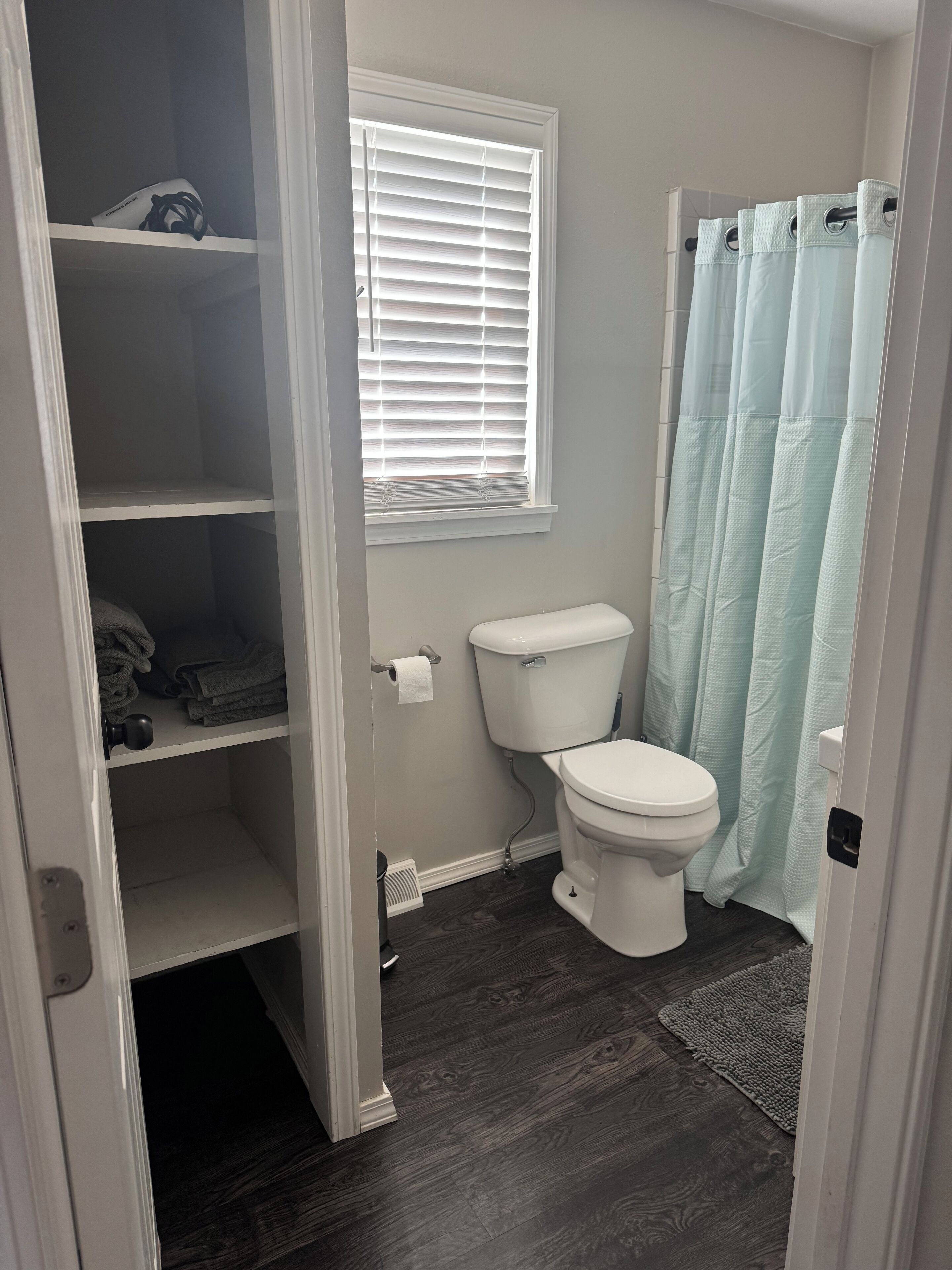 Une salle de bain avec des toilettes, une fenêtre avec des stores, un rideau de douche et un plancher en bois.