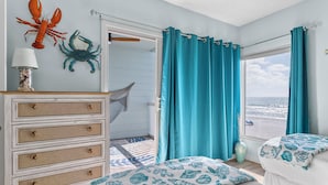 A bedroom with a view of the beach, a dresser with drawers, a bed with a blue and white patterned bedspread, and a wall with a lobster and crab decoration. - Seaside Beach House 201 · 2 Full Baths · Gulf Front (Holmes Beach)