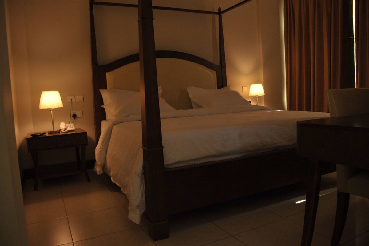 A four-poster bed with a wooden frame and white bedding, two bedside lamps, and a chair to the right.