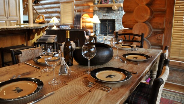 Une table à manger dressée avec des assiettes, des verres et des couverts, entourée de chaises en bois et agrémentée d’une cheminée en pierre, le tout dans l’intérieur d’un chalet en rondins.