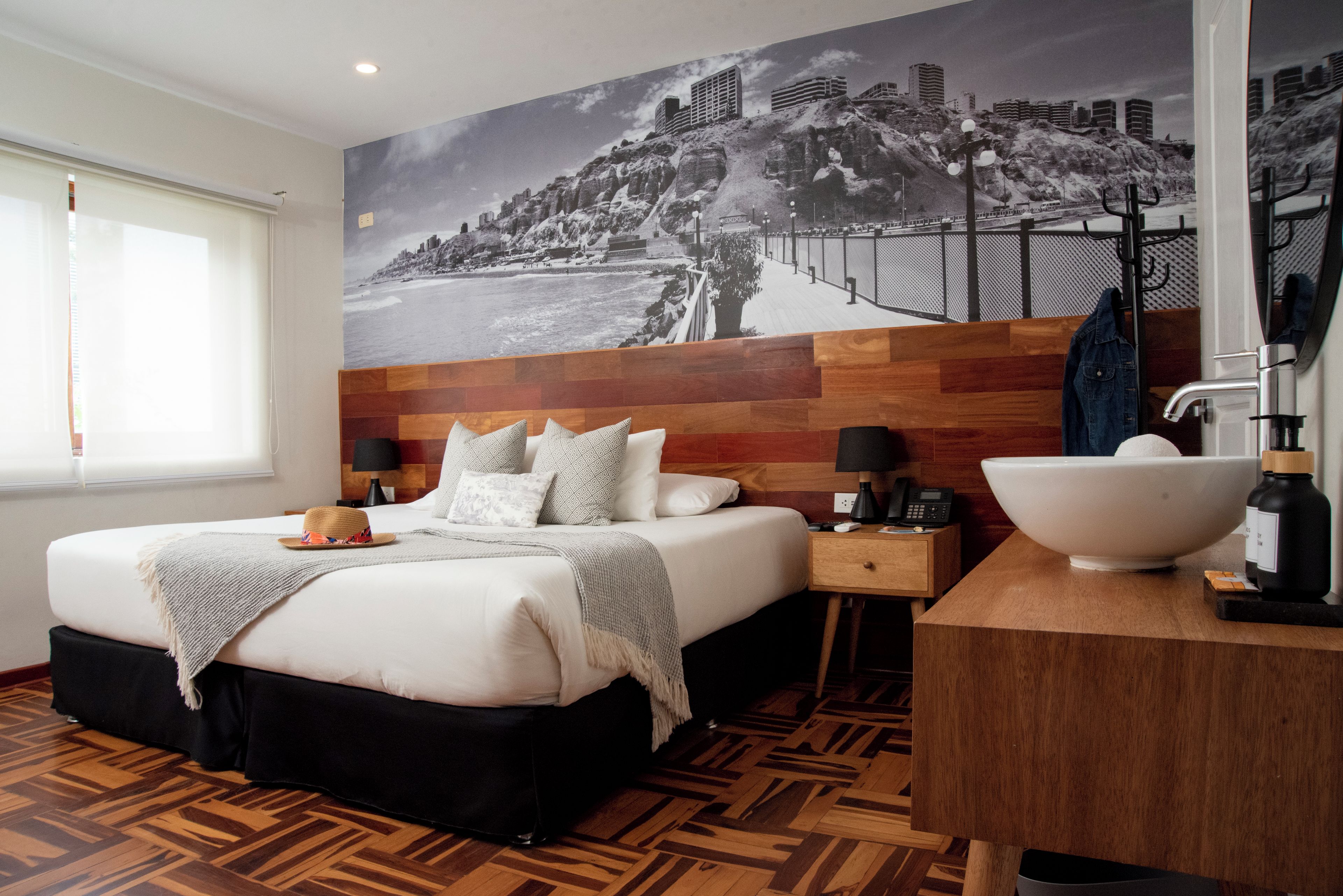 Habitació d'hotel amb llit, una petitetta, finestra gran, mirall fixat a la paret, sòl de fusta i mural que representa un paisatge urbà.