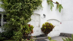 Un bâtiment blanc avec deux portes blanches, un petit jardin agrémenté de diverses plantes et un escalier en pierre menant à l’entrée.
