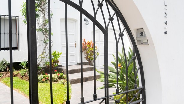Un bâtiment blanc avec un portail en fer noir, une porte blanche et un petit jardin agrémenté de plantes et de fleurs.