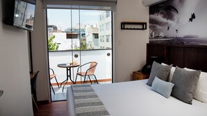 Une chambre d’hôtel avec un lit, un téléviseur fixé au mur, un balcon avec une table et des chaises, et une fresque représentant un paysage urbain sur le mur.