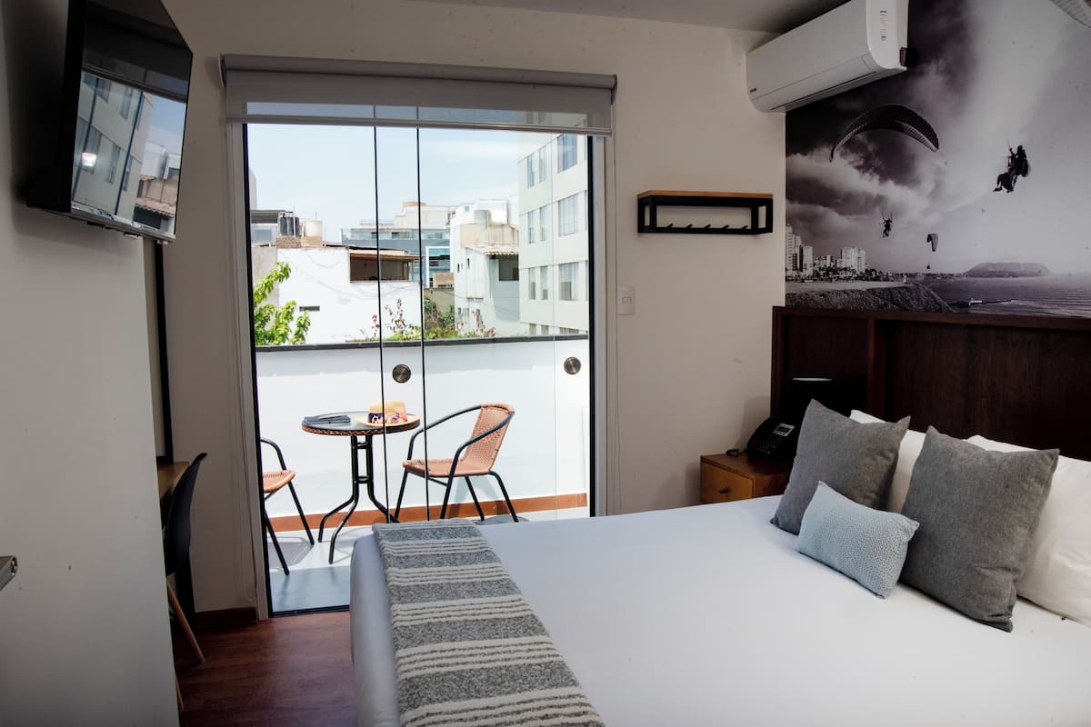 Una habitación de hotel con una cama, un televisor montado en la pared, un balcón con mesa y sillas y un mural de paisaje urbano en la pared.