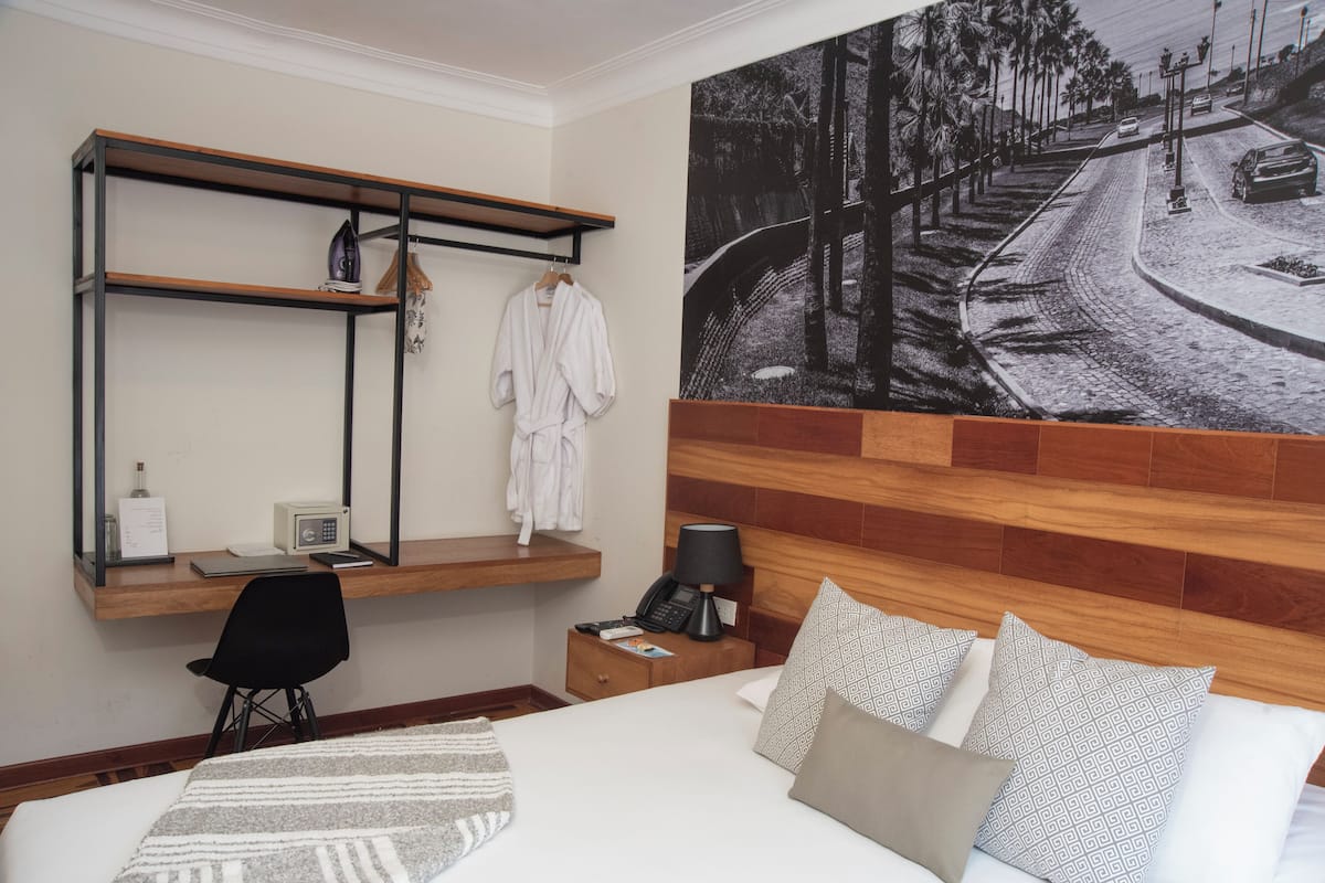 Habitación de hotel con cama, escritorio con silla, mesita de noche, estante en la pared con una bata y una fotografía grande de una escena callejera.
