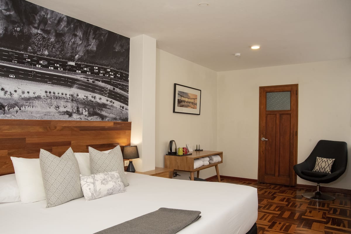 Habitación de hotel con una cama grande, cabecera de madera, un escritorio con silla, puerta de madera y una fotografía grande en blanco y negro en la pared.