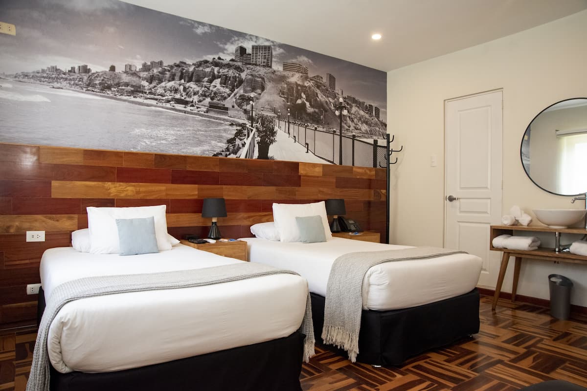 Habitación de hotel con dos camas, un mural grande en la pared, cabecero de madera, mesita de noche con lámpara, espejo y un estante pequeño con toallas.
