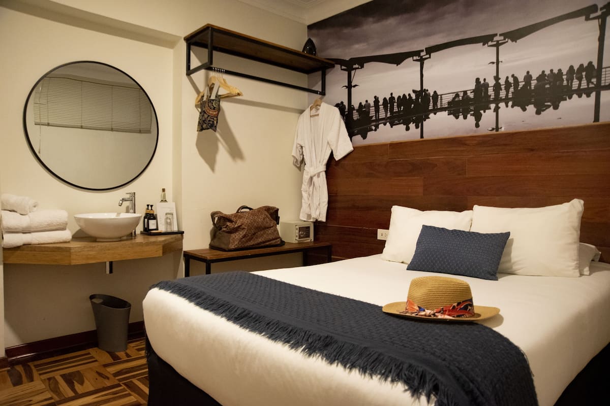 Una habitación de hotel con una cama grande, un espejo redondo, un cabecero de madera, una mesita de noche con una lámpara, un perchero para toallas, piso de madera y un mural en la pared.