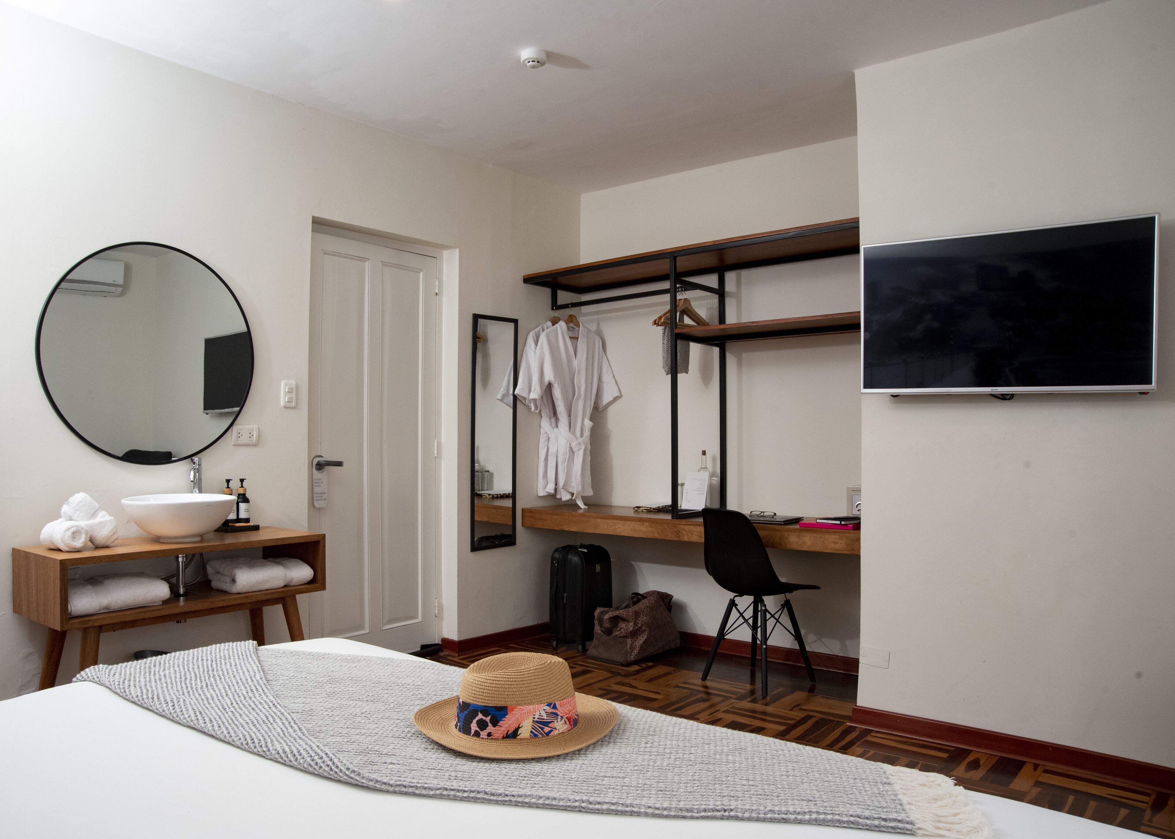 A hotel room with a bed, a round mirror, a wooden vanity with a sink, a robe rack, a desk with a chair, and a flat-screen TV.
