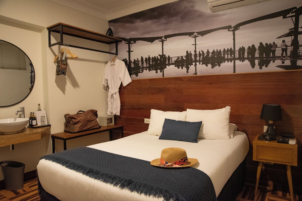 A hotel room with a wooden headboard, a large bed with white and blue pillows, a wooden nightstand with a lamp, a wooden shelf with a robe and bag, a round mirror, and a wall mural.