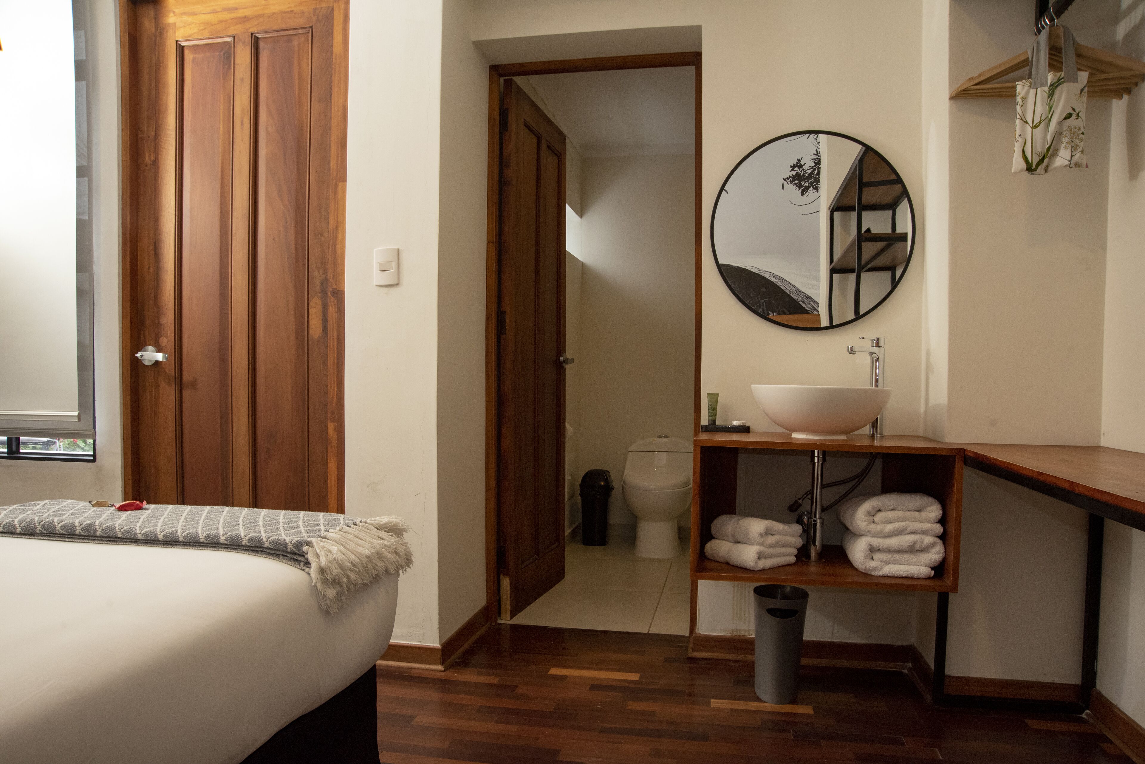 Un dormitorio con un armario de madera, una cama con manta gris, un espejo redondo, un estante de madera con toallas y un lavabo blanco.