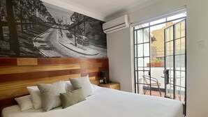 Une chambre d’hôtel avec un grand lit, une tête de lit en bois, des coussins à motifs, une table de chevet, une lampe et une fresque murale représentant une scène de rue.