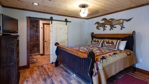 A bedroom with a wooden bed, a mounted TV, a horse decoration on the wall, and a rustic wooden ceiling.
