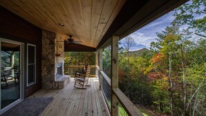 A wooden deck with a stone fireplace, outdoor seating, and a scenic view of autumn foliage.