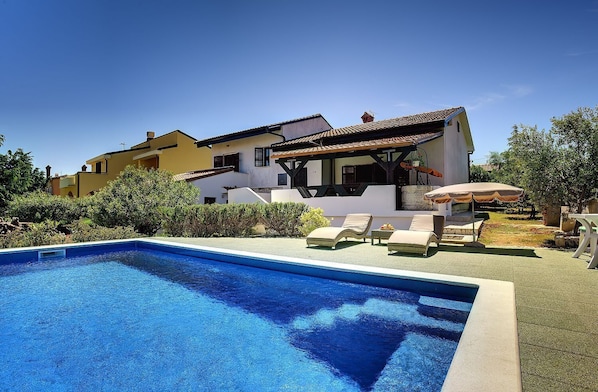 Una piscina con sillas de descanso y una zona de estar cubierta, rodeada de un jardín ajardinado y una casa de dos pisos con tejado de baldosas.