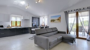 Una cocina moderna con gabinetes blancos, una zona de comedor con mesa y sillas, un sofá gris y ventanas grandes con cortinas translúcidas.
