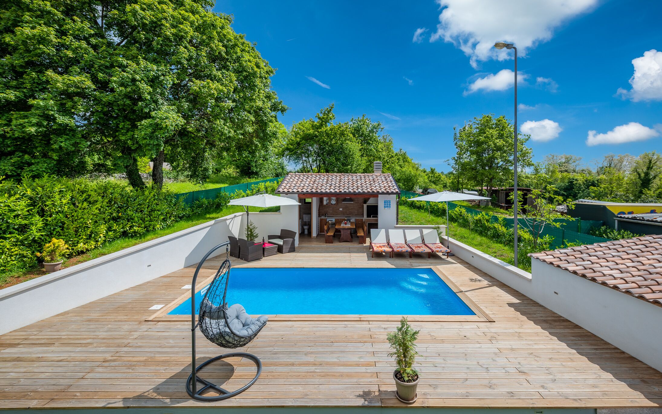 A wooden deck with a swimming pool, outdoor seating, and a small building with a tiled roof.
