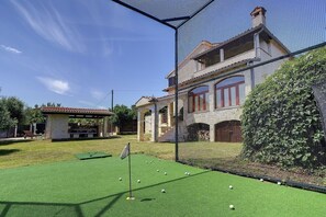 Zona de práctica de golf con césped artificial, una cerca de malla y un edificio con techo de tejas al fondo.