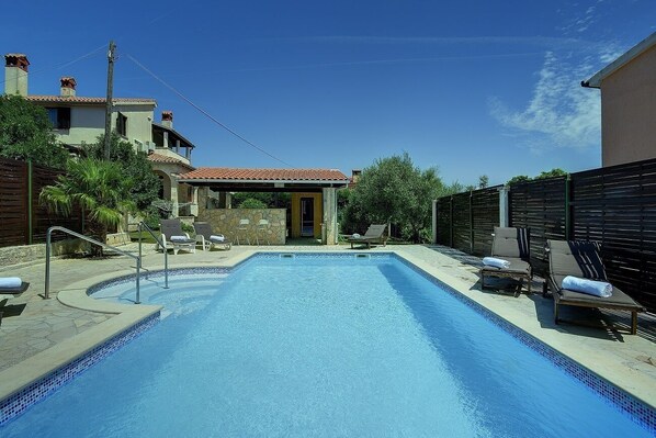 Una piscina rectangular con borde de azulejos, rodeada de sillas de descanso y un muro de piedra.
