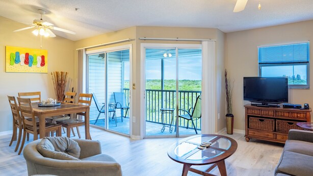 A spacious living room with a dining area, a balcony with a view, and a television on a wooden cabinet. - Sandy Pointe 210 · Mangrove & Bay View · Pool (Holmes Beach)