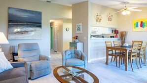 A spacious living room with a large painting, a dining area with wooden furniture, and a kitchen with white cabinets. - Sandy Pointe 210 · Mangrove & Bay View · Pool (Holmes Beach)