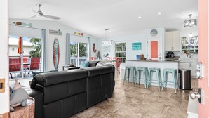 A spacious living area with a black leather sofa, a dining table with chairs, and a kitchen area with a bar counter and stools. - Sirena Del Mar | Newly Remodeled, Steps to Beach & Private Pool (Holmes Beach)