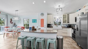 A modern kitchen with a central island, bar stools, a refrigerator, and a dining area with chairs and a table. - Sirena Del Mar | Newly Remodeled, Steps to Beach & Private Pool (Holmes Beach)