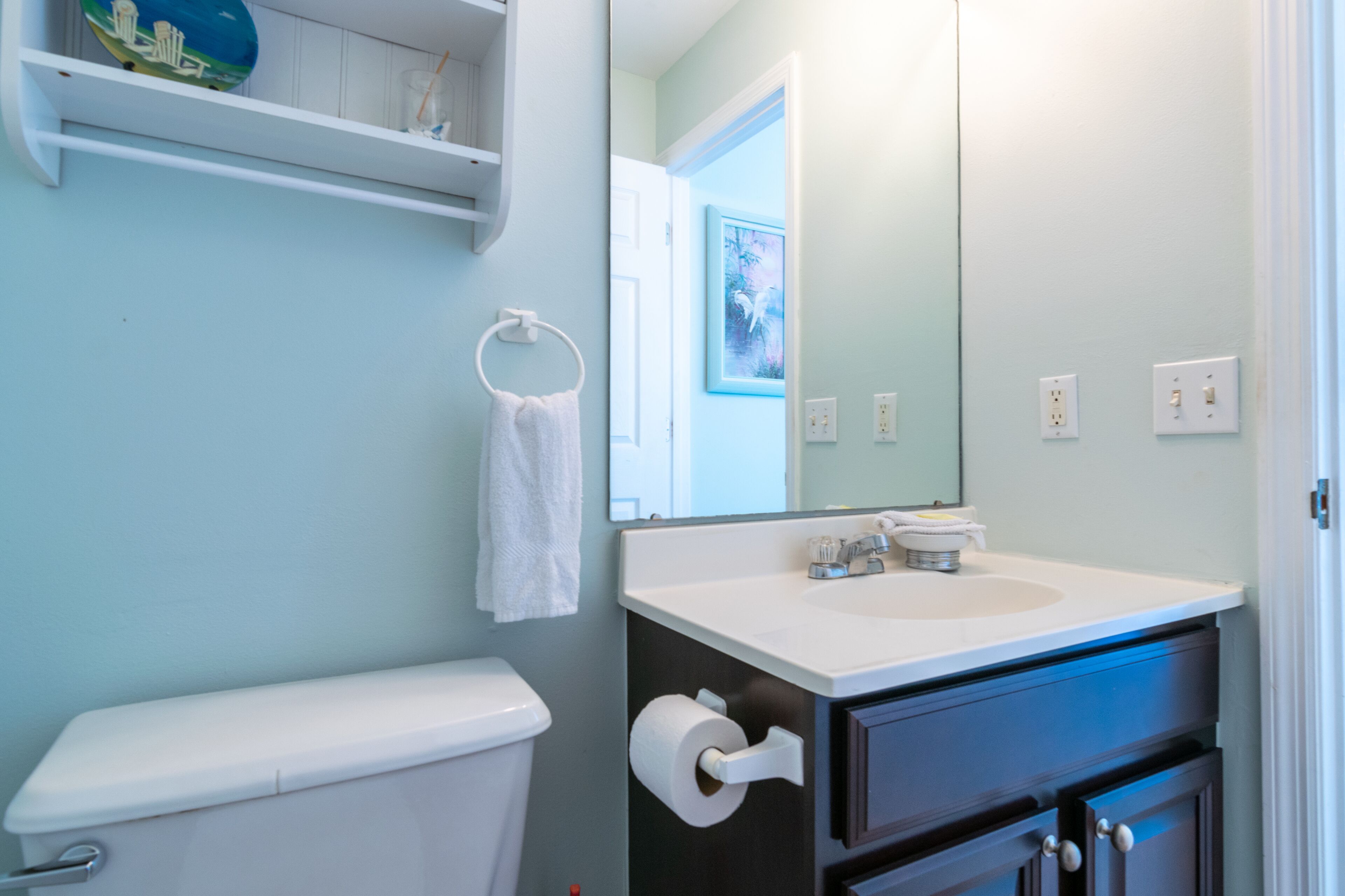 Un baño con inodoro, lavamanos, toallero, espejo y repisa con objetos decorativos.