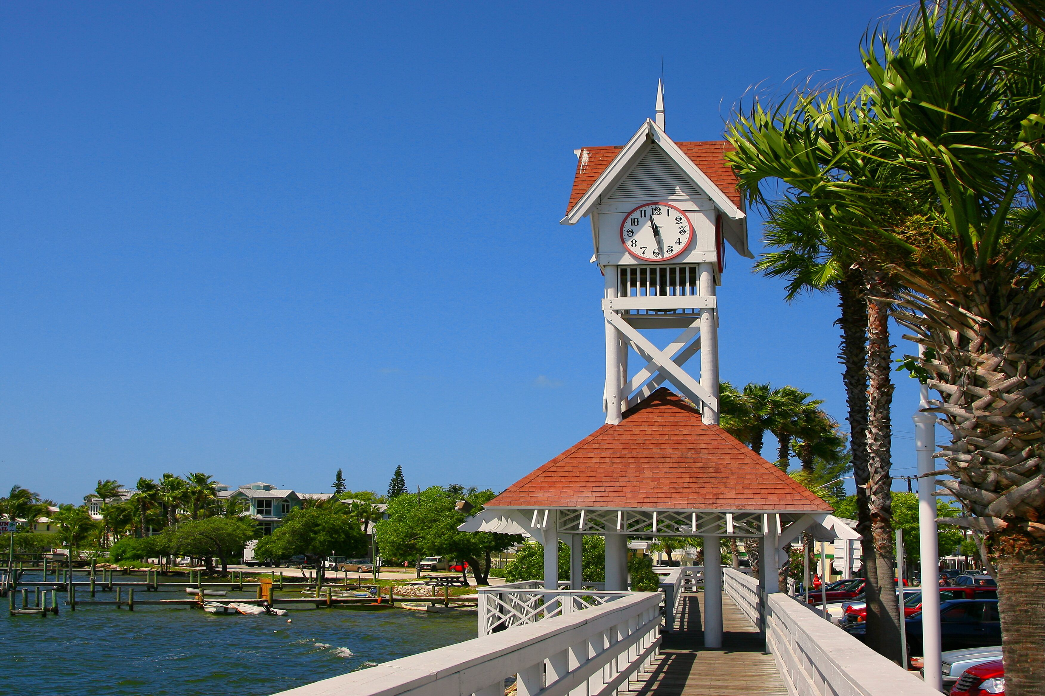 A clock tower with a red roof and white walls, overlooking a waterfront with boats and palm trees.