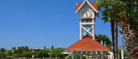A clock tower with a red roof and white walls, overlooking a waterfront with boats and palm trees.