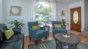 Una sala de estar con dos sillones azules, un sofá de cuero oscuro, una mesa redonda de centro y un ventanal con cortinas.