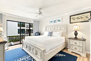 A bedroom with a large bed, a ceiling fan, a nightstand with a lamp, and a view of the outside balcony.