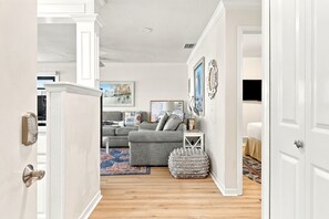 Una sala de estar con un sofá gris, una mesa de centro y una alfombra. Se observa una puerta blanca a la izquierda y una pared blanca con un marco de cuadro.