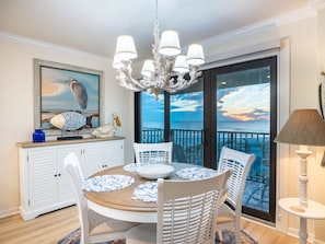 A dining area with a round table, white chairs, a chandelier, and a view of the ocean through a large glass door. - Waters Edge 110S · Direct Gulf Balcony · 1-Month Rental (Holmes Beach)