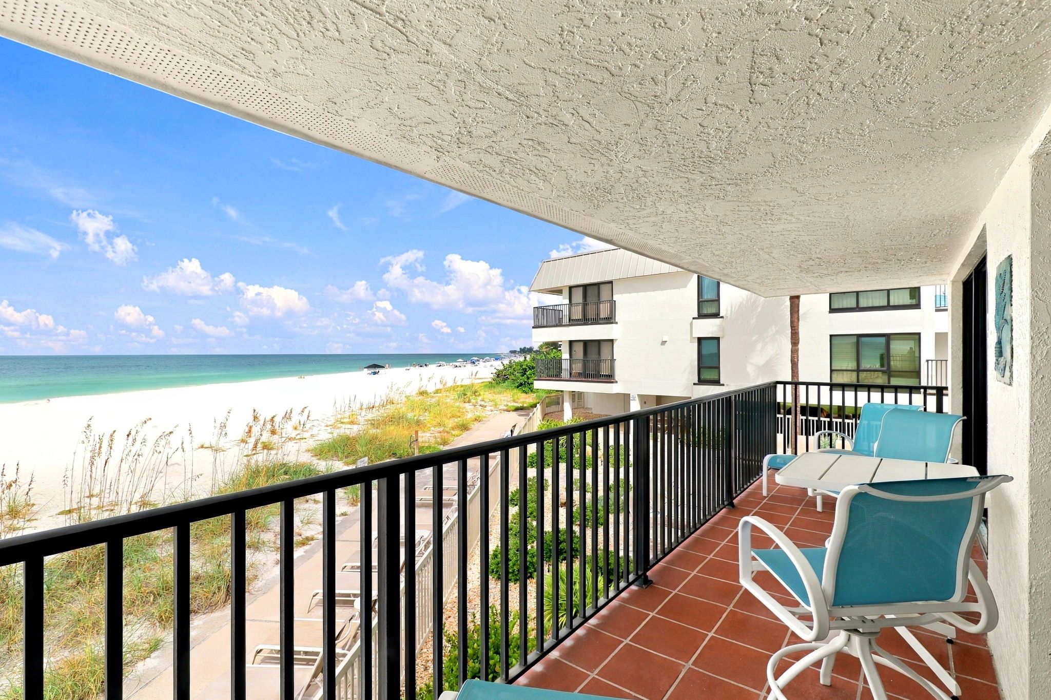 A balcony with a view of the beach, a dining area with chairs, and a building with multiple floors.