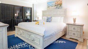 A bedroom with a large bed, a ceiling fan, a painting, a nightstand, and a blue rug.