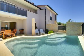 Una casa moderna de dos pisos con piscina, mobiliario exterior y un muro de piedra.