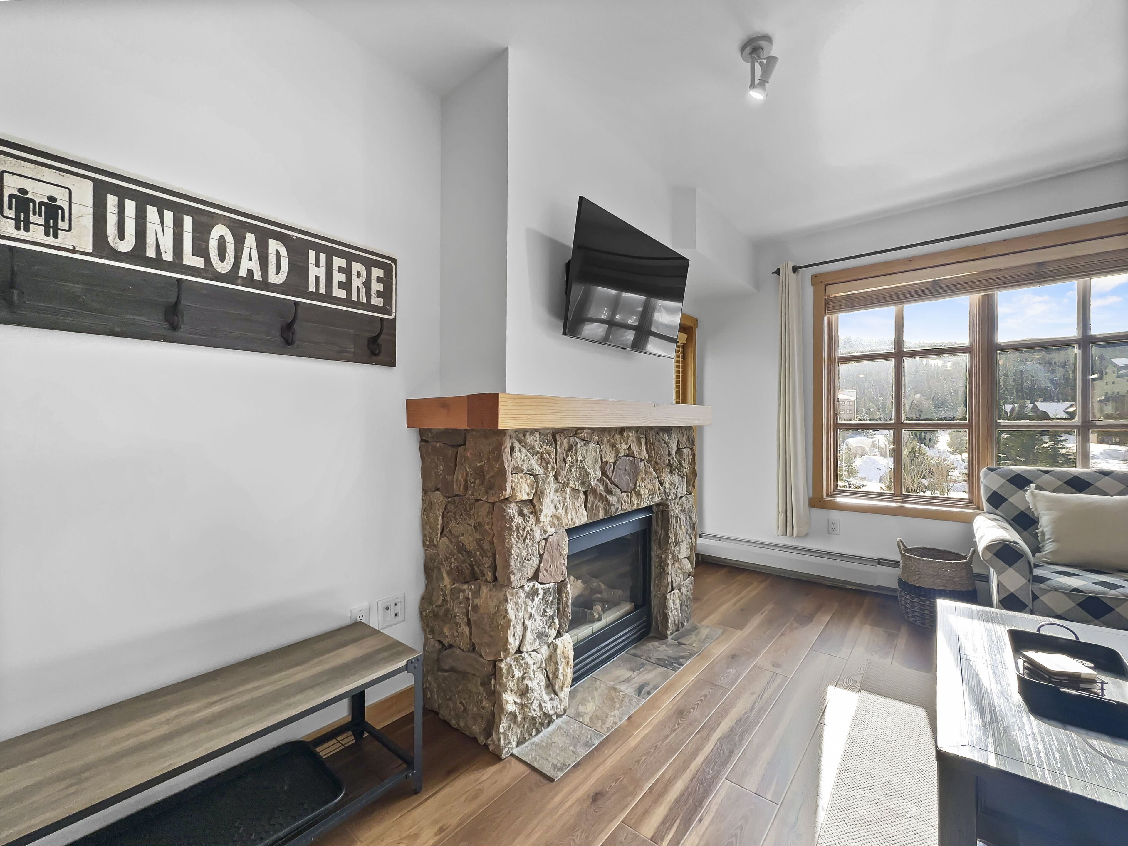 A living room with a stone fireplace, a mounted television, and a large window with a view of a snowy landscape.